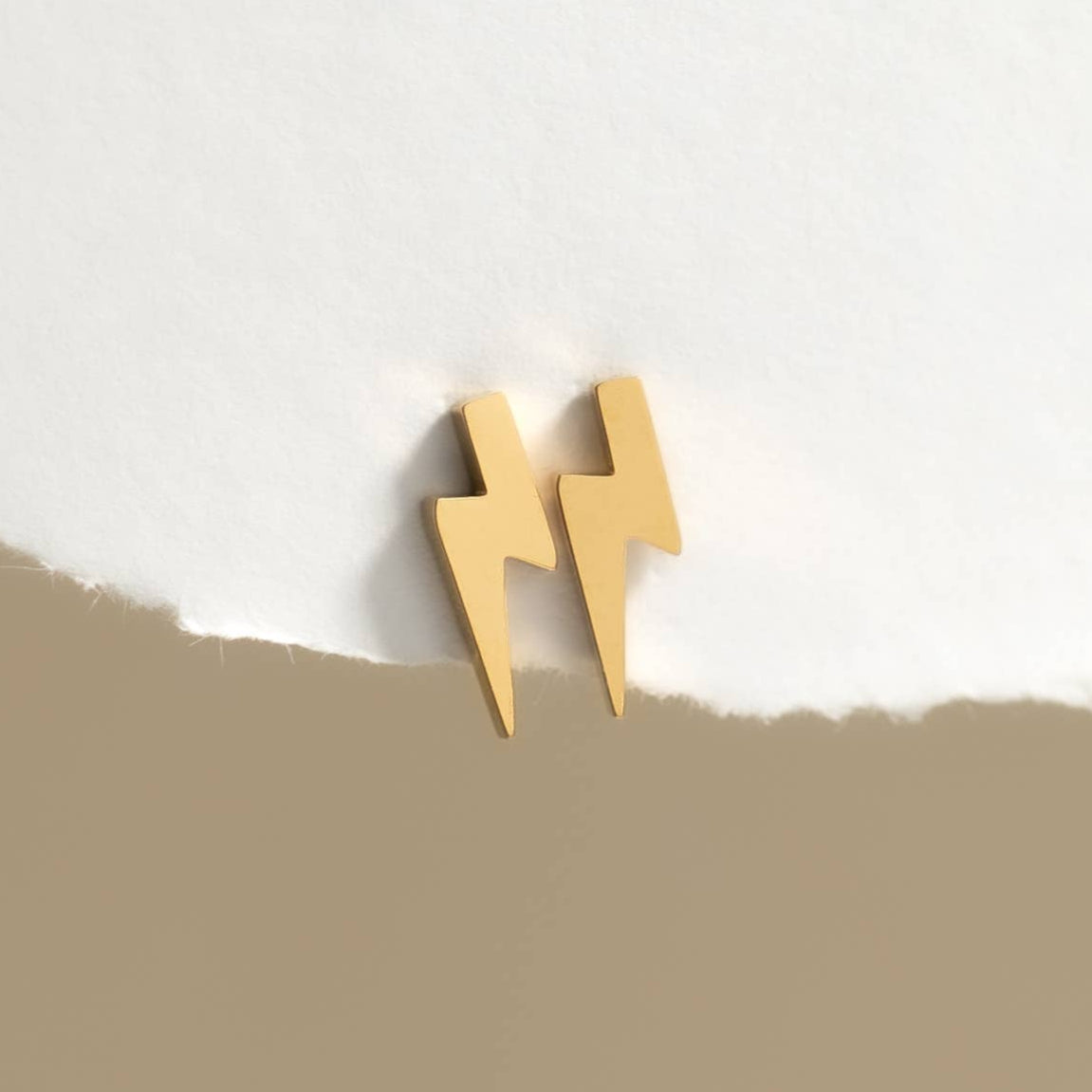 Gold lightning bolt-shaped earrings on a white background with a beige strip at the bottom.
